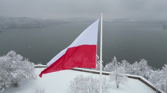 Polish flag gracefully flutters in the cold winter breeze against a serene lake and snow-covered forest. Flag of Poland waves gently in the winter wind with tranquil lake and snowy trees at background