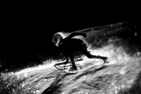 Helmeted skeleton athlete pushing sled on snowy slope with motion trails and grain effect, monochrome. Winter sports media, racing equipment concept.