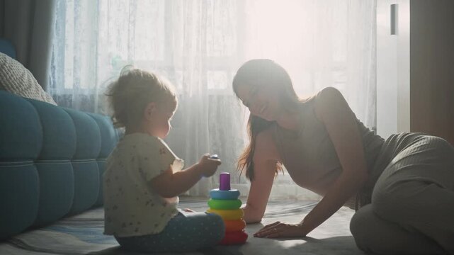 Soft sunlight fills the room. Woman and child play with colorful stacking toys. They sit close, hands joined, smiling gently. Warmth and joy fill the quiet moment. Perfect for family, parenting