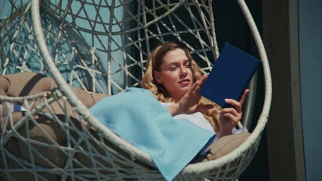 Woman relaxes in hanging chair, reading book. Soft sunlight falls on her peaceful expression. Blue blanket rests beside her, adding comfort. Ideal for scenes of calm, leisure, or cozy reading moments