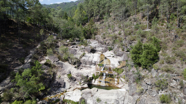 Aerial view of Cascatas de Fecha de Barjas waterfall cascading over rocky terrain surrounded by lush pine forest in Salamonde, Braga, Portugal.