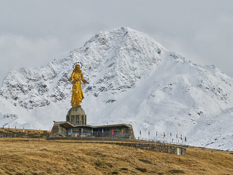 Aerial view of the golden Madonna di Madesimo statue standing on a stone pedestal with a row of flags against a backdrop of snow-covered mountains in Lombardia, Italy.