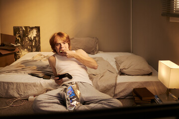 Caucasian young adult male sitting on floor playing video games with joystick while eating snacks. Useful for gaming advertising and youth lifestyle marketing campaigns © DragonImages