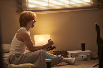 Caucasian teenage boy sitting on floor playing video games with joystick controller in bedroom. Concept representing youth entertainment, gaming lifestyle, and digital leisure marketing © DragonImages