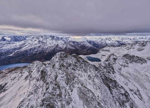 Aerial view of Engadin mountains with snow-covered peaks, alpine lakes, and dramatic cloudy sky in Grisons, Switzerland.