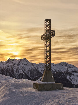 Aerial view of a metal summit cross standing on a snow-covered mountain peak against a golden sunset sky with distant snow-capped peaks in Lombardy, Italy.