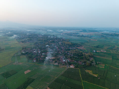 Aerial view of a village surrounded by lush green cornfields and agricultural land with thin smoke rising from the houses in Kecamatan Kembang, Jawa Tengah, Indonesia.
