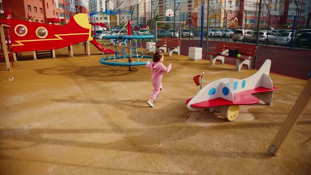 Child runs joyfully past airplane ride at playground. Bright colors frame cheerful playtime. Sunlit sand surrounds colorful structures. Perfect for children's activity or summer scenes
