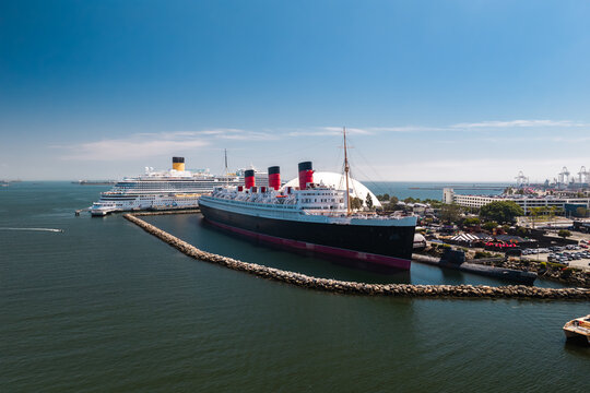 Aerial view of the historic Queen Mary ocean liner and a modern cruise ship docked along the coast in Long Beach harbor, California, beside a rock breakwater on a sunny day.