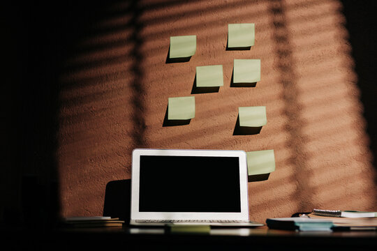 Laptop, sticky notes, notebooks on minimal home office desk, sunlight shadows, copy space