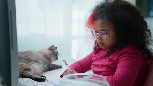 Young girl writes at desk with cat nearby. She pets the cat gently while focused on her work. Child wears glasses and pink top, showing calm concentration. Cat rests beside her, relaxed and content