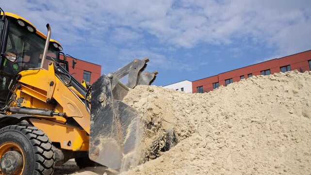 Backhoe loader pushes a mound of sand at a construction site in direct daylight, Heavy equipment moves aggregate beside new residential buildings