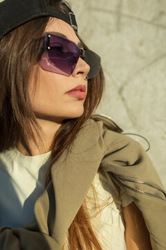 Fashion portrait of a stylish woman with a side profile, wearing unique geometric rimless sunglasses with purple lenses and a black backwards cap against a concrete wall. Street style look.