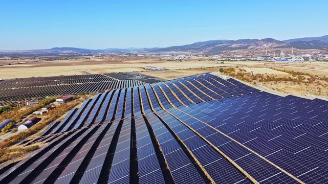 Rows of solar panels are arranged in neat patterns across a wide open space in a rural area. The mountains can be seen in the distance.
