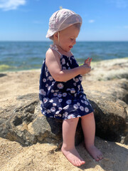  Side view of a little toddler girl sitting on a large rock on a sandy sea beach during a summer vacation. © Irina