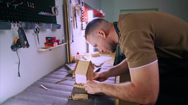 Man focuses intently on crafting a small wooden house. Tools hang neatly on walls behind him. Workshop is tidy and well-lit. Crafting shows dedication and care. Perfect for DIY, home