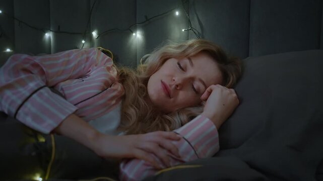 Soft lights glow above a sleeping woman. She rests peacefully in pink striped pajamas. Her arms lie gently across her chest. Dark bedding frames her tranquil expression