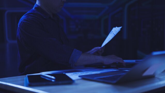 Asian programmer analyzes financial paper document, enters server data late night. Cyber security audit, corporate database network maintenance, overtime software engineering troubleshooting.
