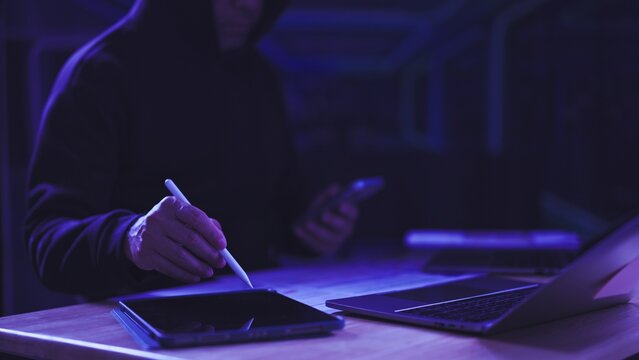 Hooded hacker works on digital tablet near laptop computer in dark neon room. Cybercrime threat prevention, data breach investigation, information security breach, network espionage attack.