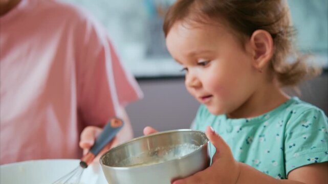 Toddler watches with curiosity while adult whisks batter. Child holds bowl, eyes fixed on mixing. Mom guides child through kitchen activity. Perfect for family, cooking, parenting
