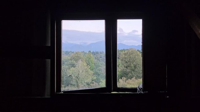 Green treetops and mountain ridges seen through a wooden window in a dark rustic interior. Horizontal view contrasts the shadowed room with daylight and a peaceful landscape beneath cloudy skies