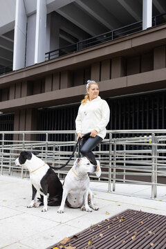 Young woman owner standing with her two pitbull walks dogs on leashes near modern city building
