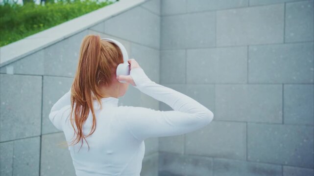 Young woman puts on white wireless headphones. She stands near gray stone wall outdoors. Her hair is tied back in a ponytail. She wears a plain white long-sleeved top