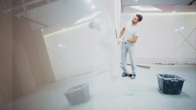 Young man smiles while painting a white wall. He stands in a bright, empty room. Two buckets sit on the floor nearby. He appears happy and focused on the task