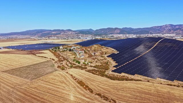 A large solar farm spans across open land near a city. It features numerous solar panels aligned in rows. Nearby, there are scattered buildings and hills in the distance under a clear sky.