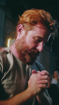 Caucasian man with red beard praying with folded hands and head tilted back in deep devotion during indoor church worship service