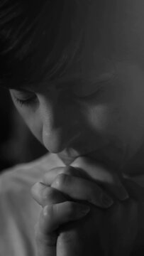 Black and white close-up of woman praying with folded hands held to chin in deep devotion and emotional faith during indoor worship