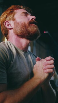 Caucasian man with red beard praying with folded hands and head tilted back in deep devotion during indoor church worship service