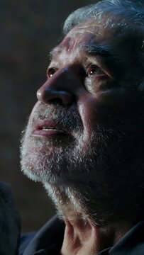 Elderly senior man praying with clasped hands looking upward with tearful emotion and hope during indoor church worship service