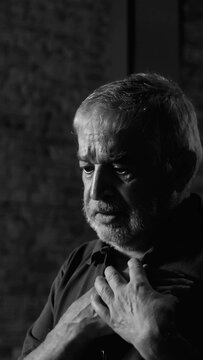 Black and white photo of senior elderly man placing hands over heart with deep emotion and sorrow during indoor church worship service