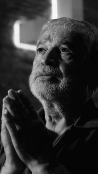 Black and white photo of senior elderly man placing hands over heart with deep emotion and sorrow during indoor church worship service