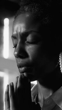 Black and white close-up of African American woman praying with folded hands and closed eyes in deep sorrow and emotional devotion indoors