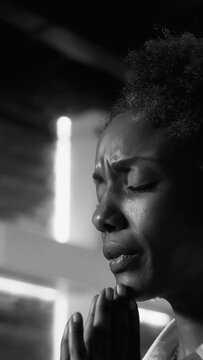 Black and white close-up of African American woman praying with folded hands and closed eyes in deep sorrow and emotional devotion indoors