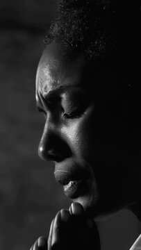 Black and white close-up of African American woman praying with folded hands and closed eyes in deep sorrow and emotional devotion indoors