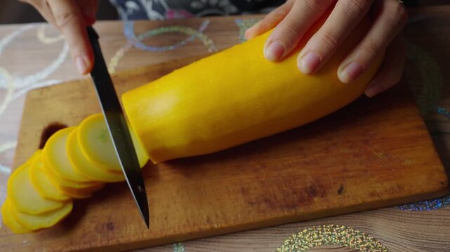 Hands slice yellow squash on wooden board. Slices pile neatly beside the main vegetable. Cutting action is steady and precise. Cooking preparation scene is calm and focused