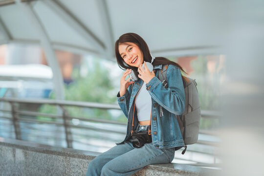Happy young Asian woman traveler sitting on walkway with backpack and camera, smiling while adjusting headphones around her neck, enjoying leisure journey, photography,