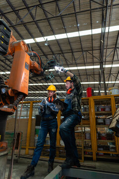 During robot inspection, two automation colleagues examine the orange arm with a flashlight inside a fenced factory cell.
