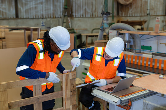 Team meeting teamwork: two factory workers inspect a wooden pallet together while one points and holds a clipboard.