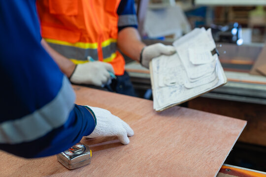 A carpenter reviews woodworking paperwork over plywood with a colleague as they examine a clipboard in the wood workshop.