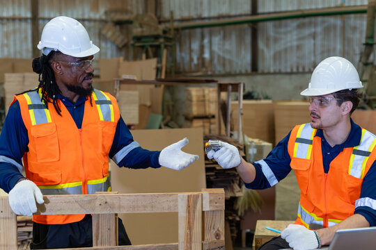 Inside woodworking premises, a carpenter discusses plywood with his colleague over a pallet and tape measure in the wood workshop.