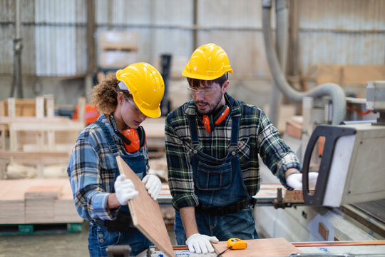 Inside woodworking premises, a carpenter holds plywood while her male colleague touches a machine in the wood workshop.
