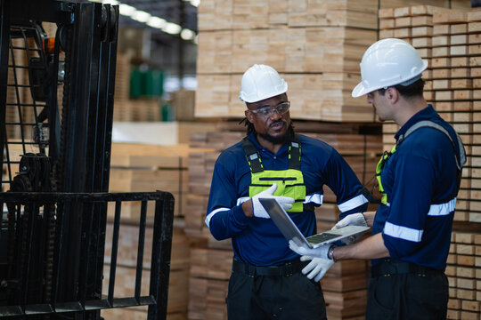 Lumber inventory is checked by two warehouse colleagues discussing information on a laptop beside tall timber stacks.