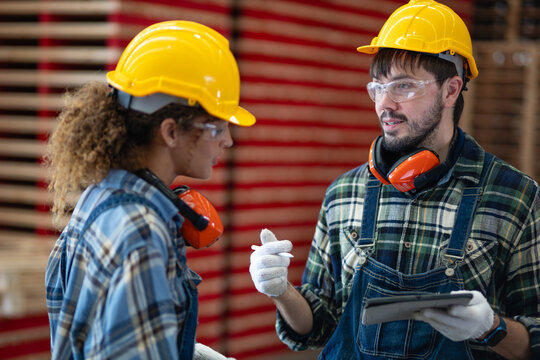 Team meeting: factory workers discuss teamwork while a man holds a tablet beside his female colleague near stacked lumber.