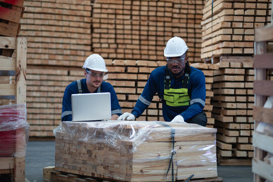 Lumber stacks surround two warehouse colleagues checking inventory as they examine a laptop resting on the wrapped wood.