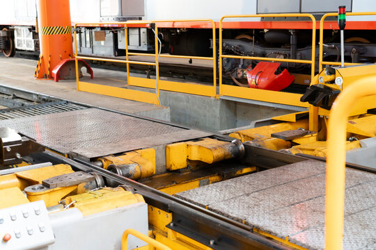 Yellow and red vintage electric railcar viewed from the front inside a railway workshop, with overhead lights, metal roof trusses, service cabinets and blue railcars nearby.