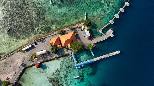 Aerial view of Karimunjawa Passenger Ferry Port with orange-roofed buildings, a long pier extending into turquoise waters, and boats docked in Karimunjawa, Central Java, Indonesia.